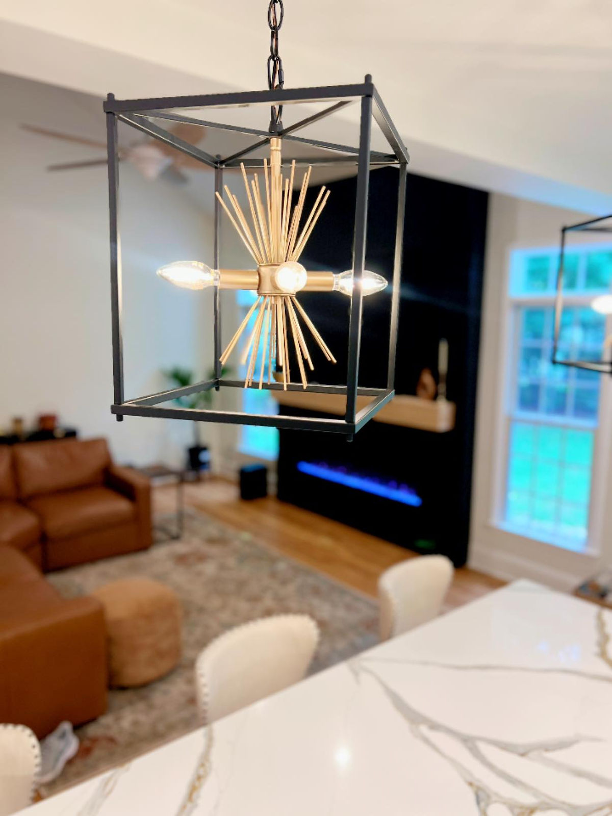 Modern dining room with a geometric chandelier. Brown couch and blue fireplace in background. Marble table in foreground. Bright and cozy.