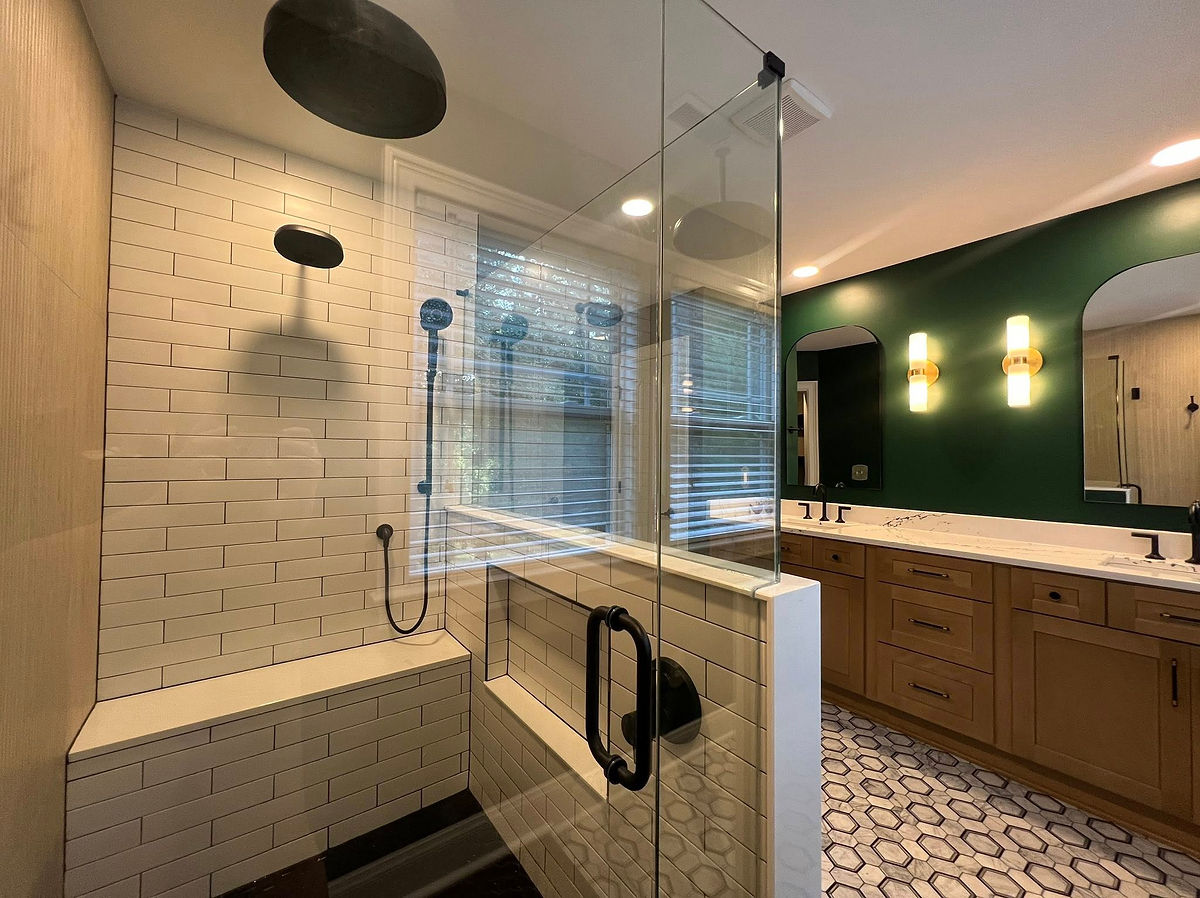 Modern bathroom with glass shower, white tiles, and black fixtures. Wooden vanity with dual sinks and green accent wall. Hexagon floor tiles finish this home renovation near Cornelius, NC.