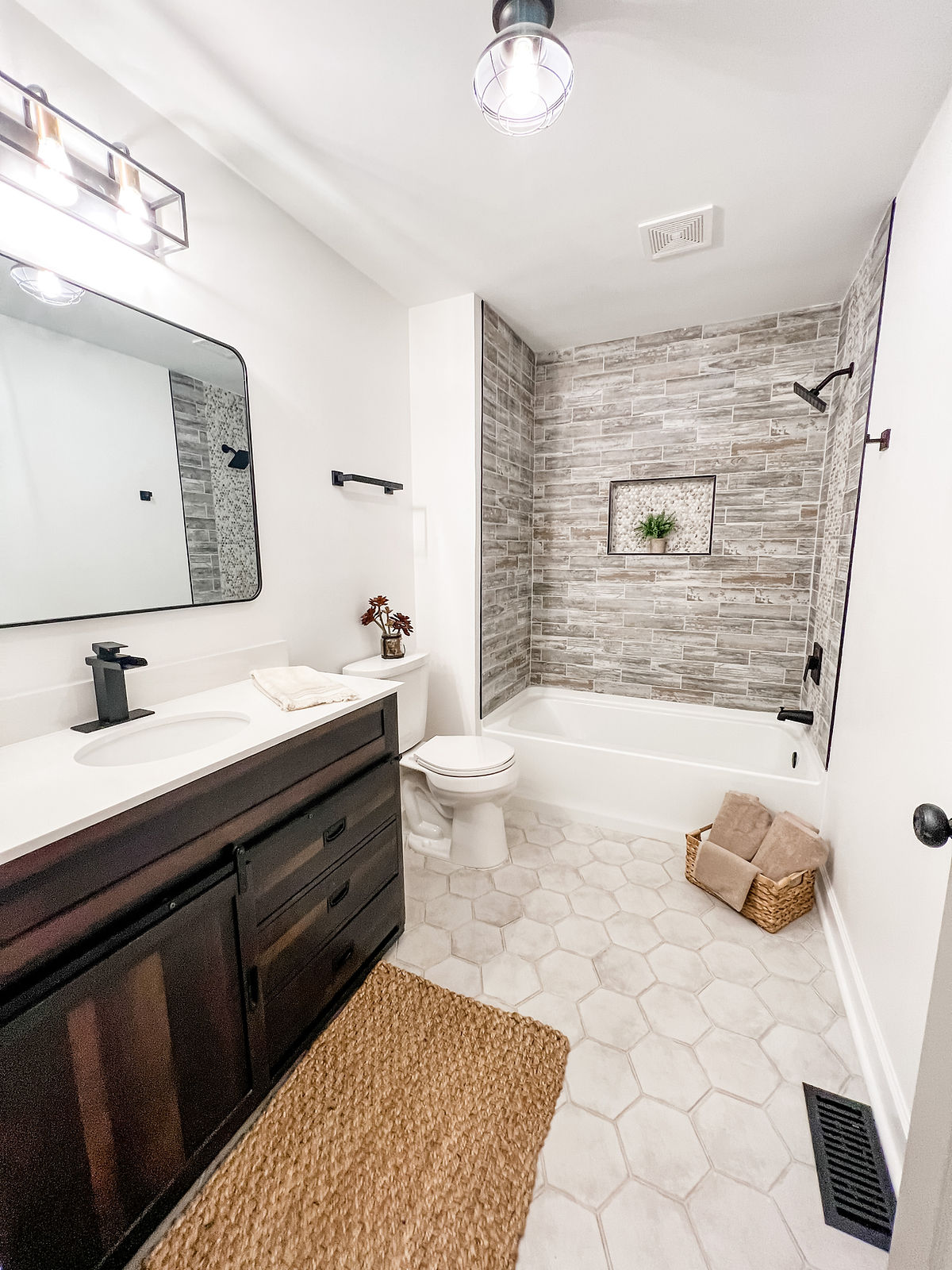 bathroom renovation shows hex tile flooring, gray tiled tub surround, a dark vanity in the foreground with white sink and counter
