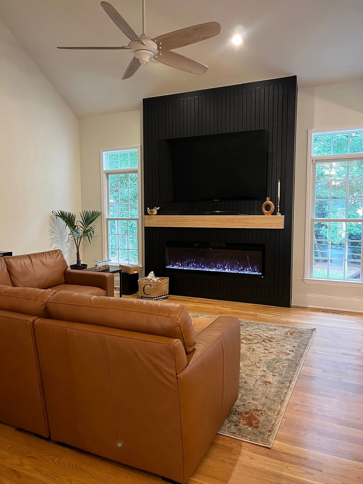 Cozy living room with brown leather sofas, black fireplace, and TV. Large windows, patterned rug, ceiling fan, and a calm atmosphere.