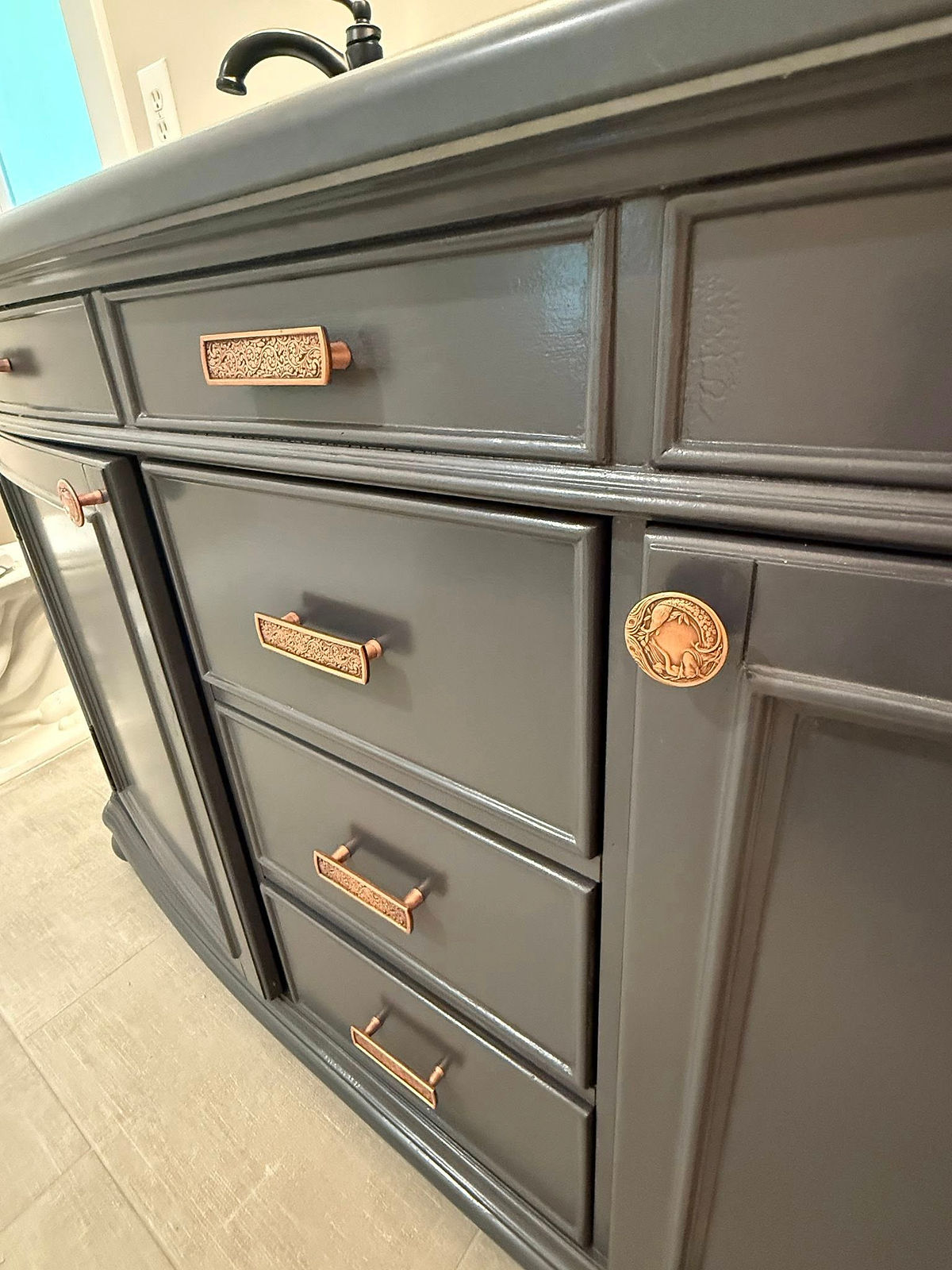 Dark gray cabinet with ornate bronze handles and a decorative medallion. Set against a tiled floor, exudes elegance and craftsmanship.