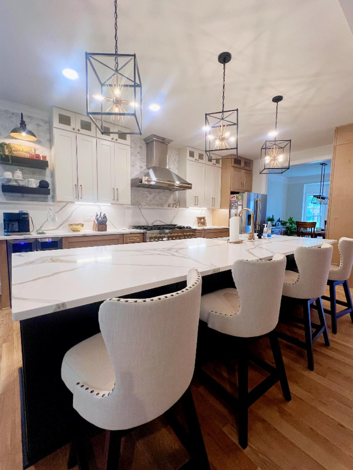 Modern kitchen with white cabinets, marble island, and four white studded chairs. Pendant lights hang overhead, creating a cozy ambiance.