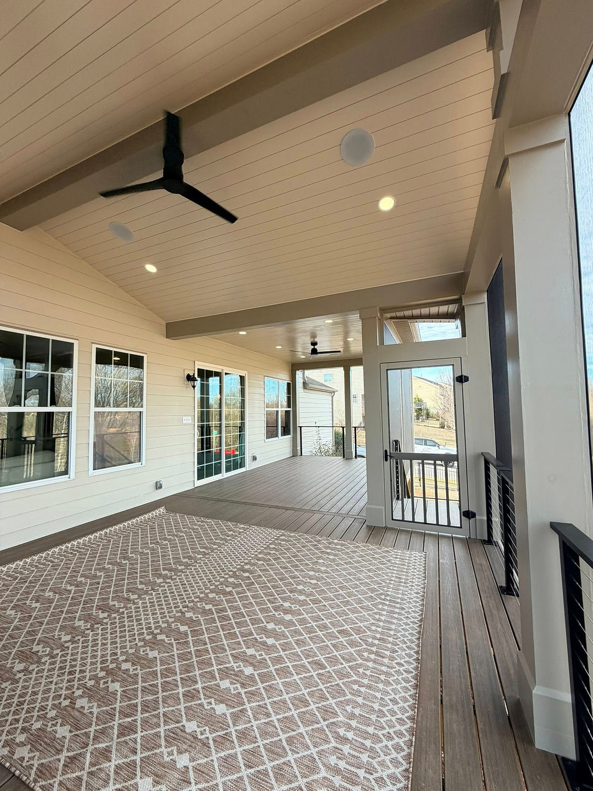 Covered patio with high ceiling, black fan, and beige geometric rug. Large windows and wood decking create an airy and modern feel.