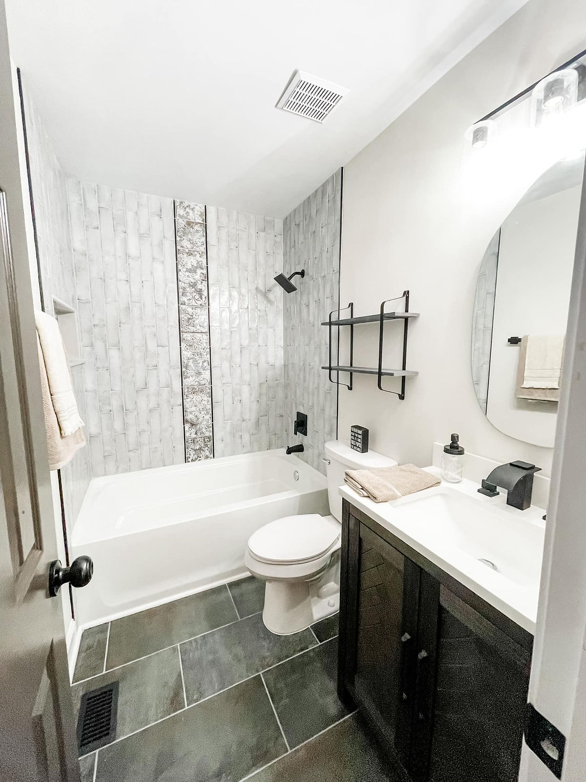 remodeled bathroom with dark tile flooring, dark vanity with white counter and sink, and decorative tile waterfall accent in the shower wall