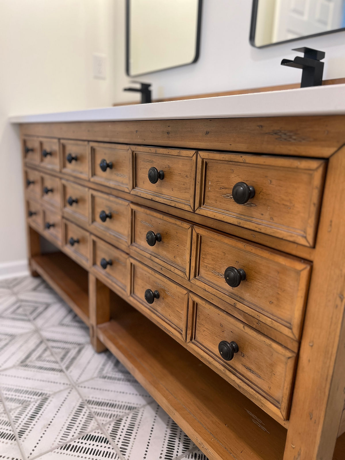 Wooden bathroom vanity with multiple drawers, black knobs, set against patterned tile floor. White countertop with two black faucets.
