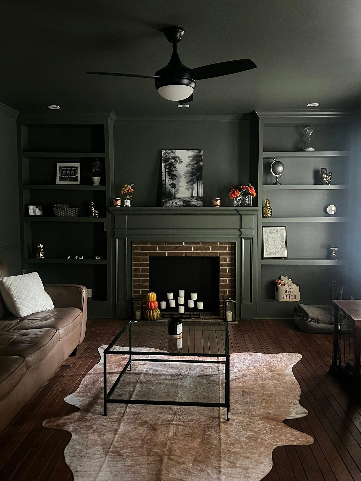 Cozy living room with a brown couch, glass table, and decor on shelves. Brick fireplace centered, with a black-and-white forest photo above. Home Renovation near Davidson, NC.