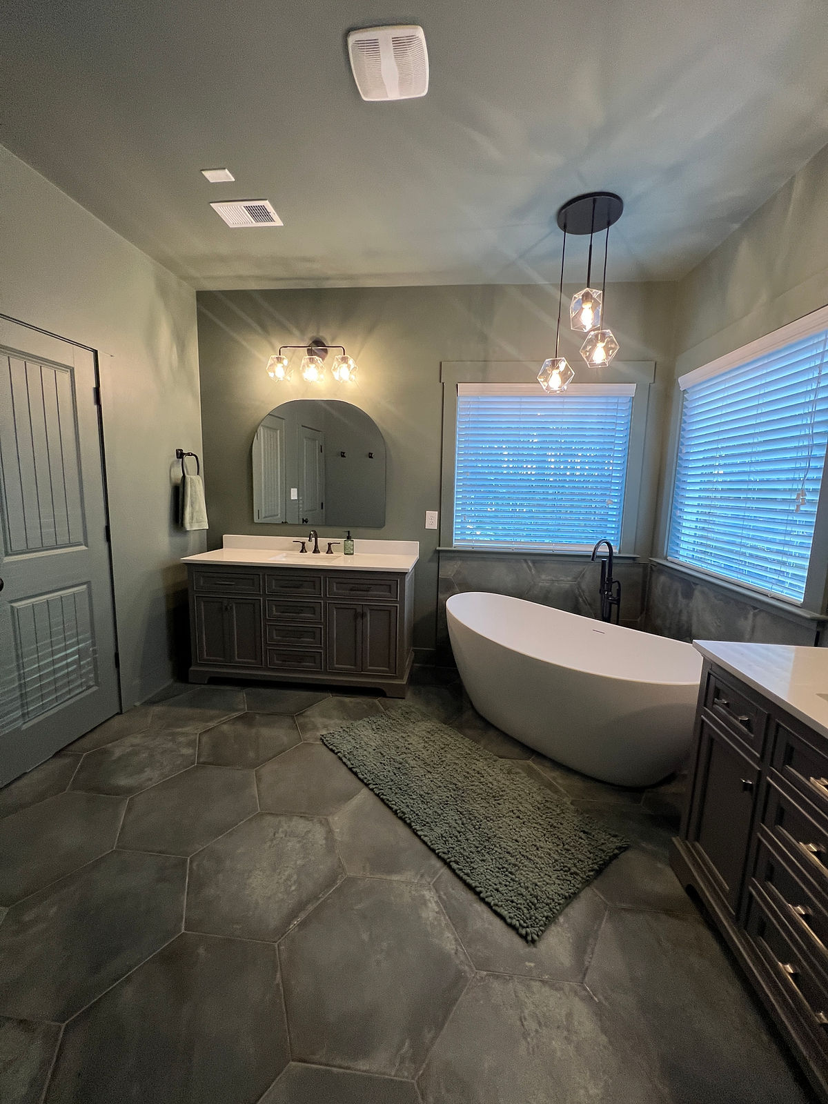 Modern bathroom with a white freestanding tub, hexagonal tiles, double vanity, large mirrors, and pendant lights. Soft green walls and blinds.