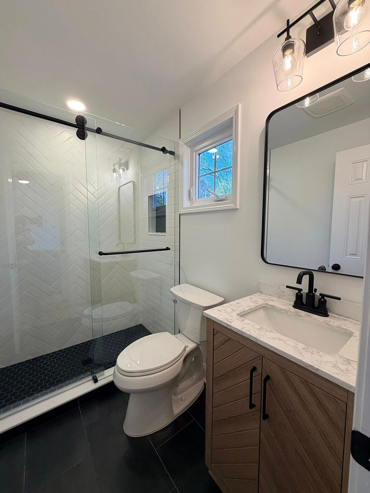 Modern bathroom with glass shower, black fixtures, chevron tile, black floor, white walls, marble vanity, wood cabinets, and a window in home renovation near Cornelius, NC.
