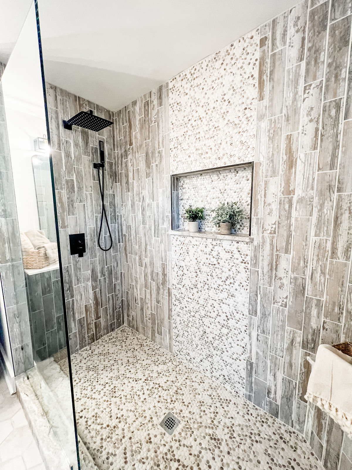 detail shot of renovated bathroom shows pebble tile waterfall accent and flooring inside a subway tiled shower stall . recessed niche holds potted plants