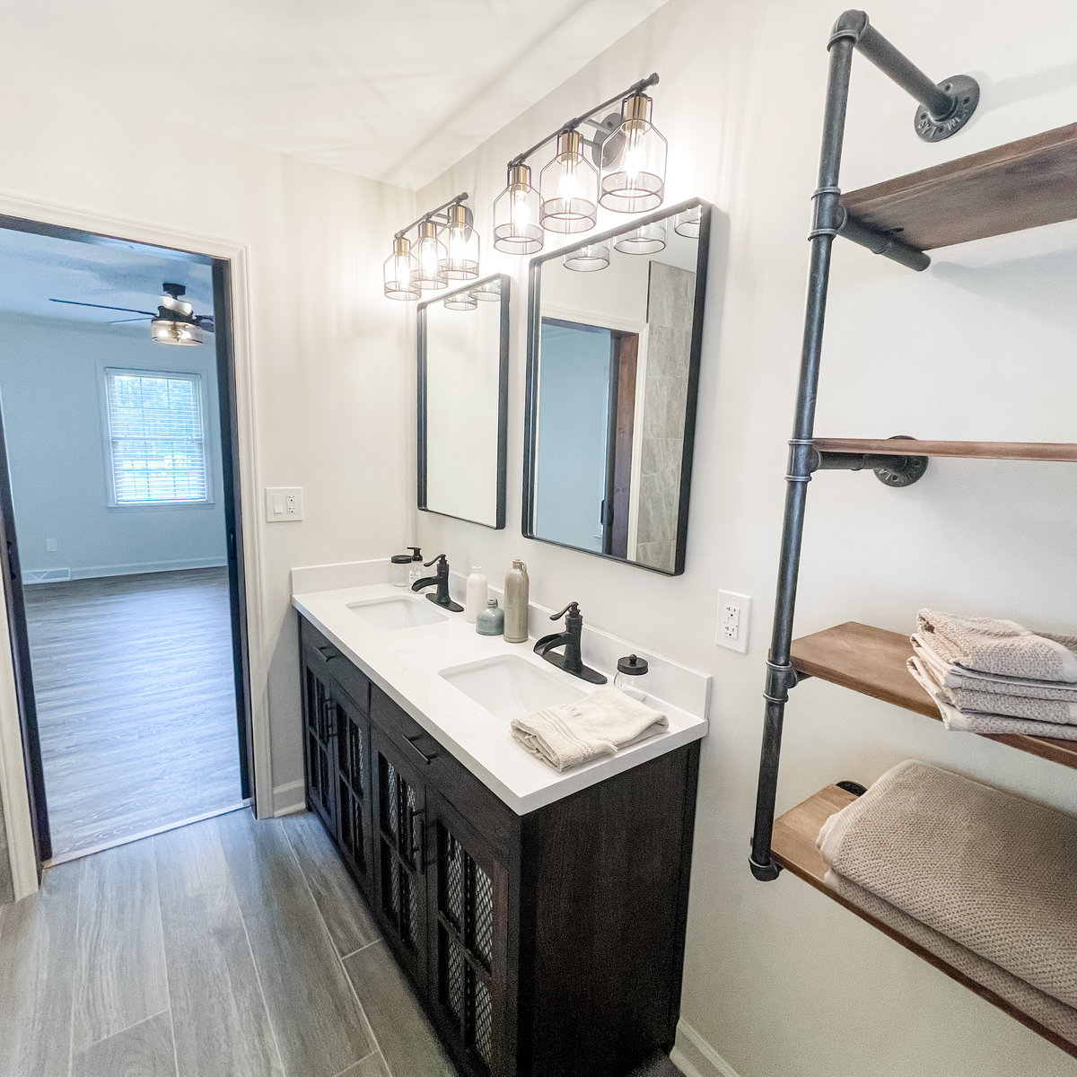 renovated bathroom with light walls, gray tile floors, dark vanity with white sink and counter, an industrial pipe style shelf is in the foreground