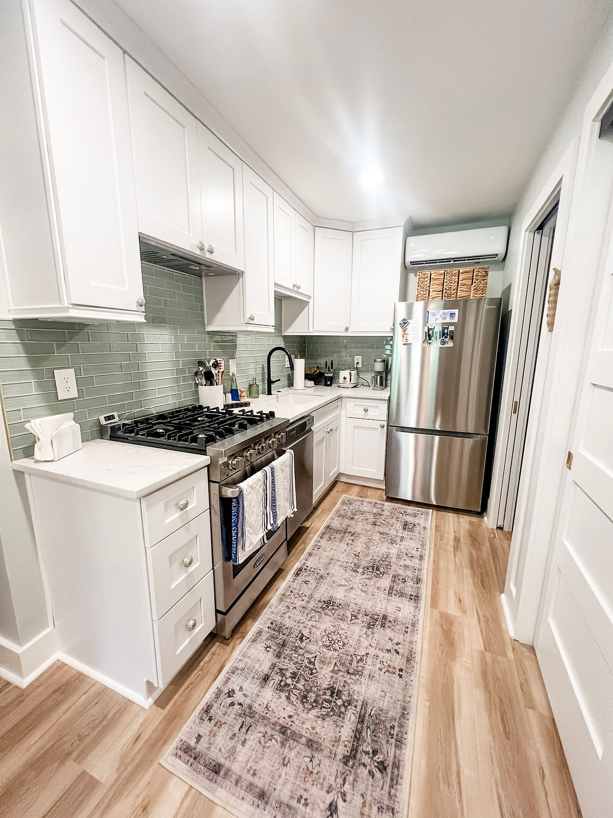 Modern kitchen with white cabinets, green tile backsplash, stainless steel appliances, and a patterned rug. Bright and tidy setting.