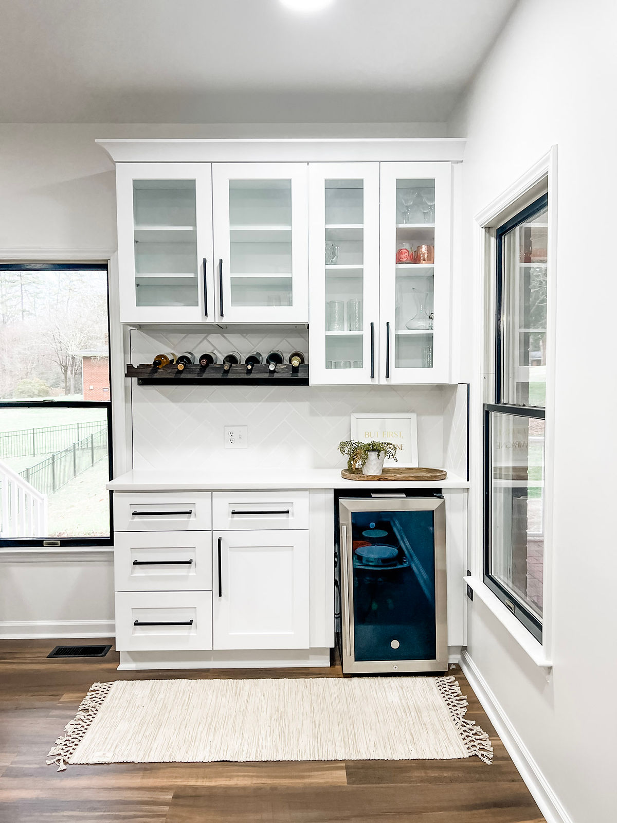 White kitchen with glass cabinets, wine rack, and mini fridge. A potted plant on the counter. Large windows and a light rug on wood floor.