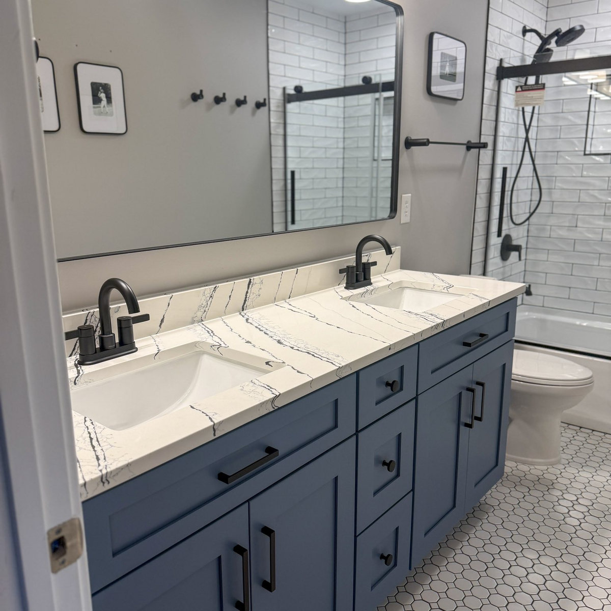 dark blue vanity with stone counters in front of a large mirror on a gray wall. tiled shower in the background