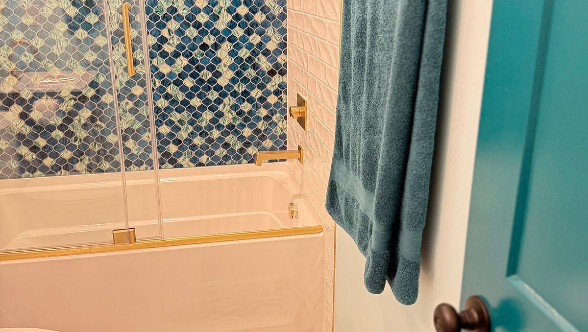 Bathroom with blue mosaic tiles, teal towel, and shower door. Features a toilet, patterned mat, and teal door, creating a fresh look.