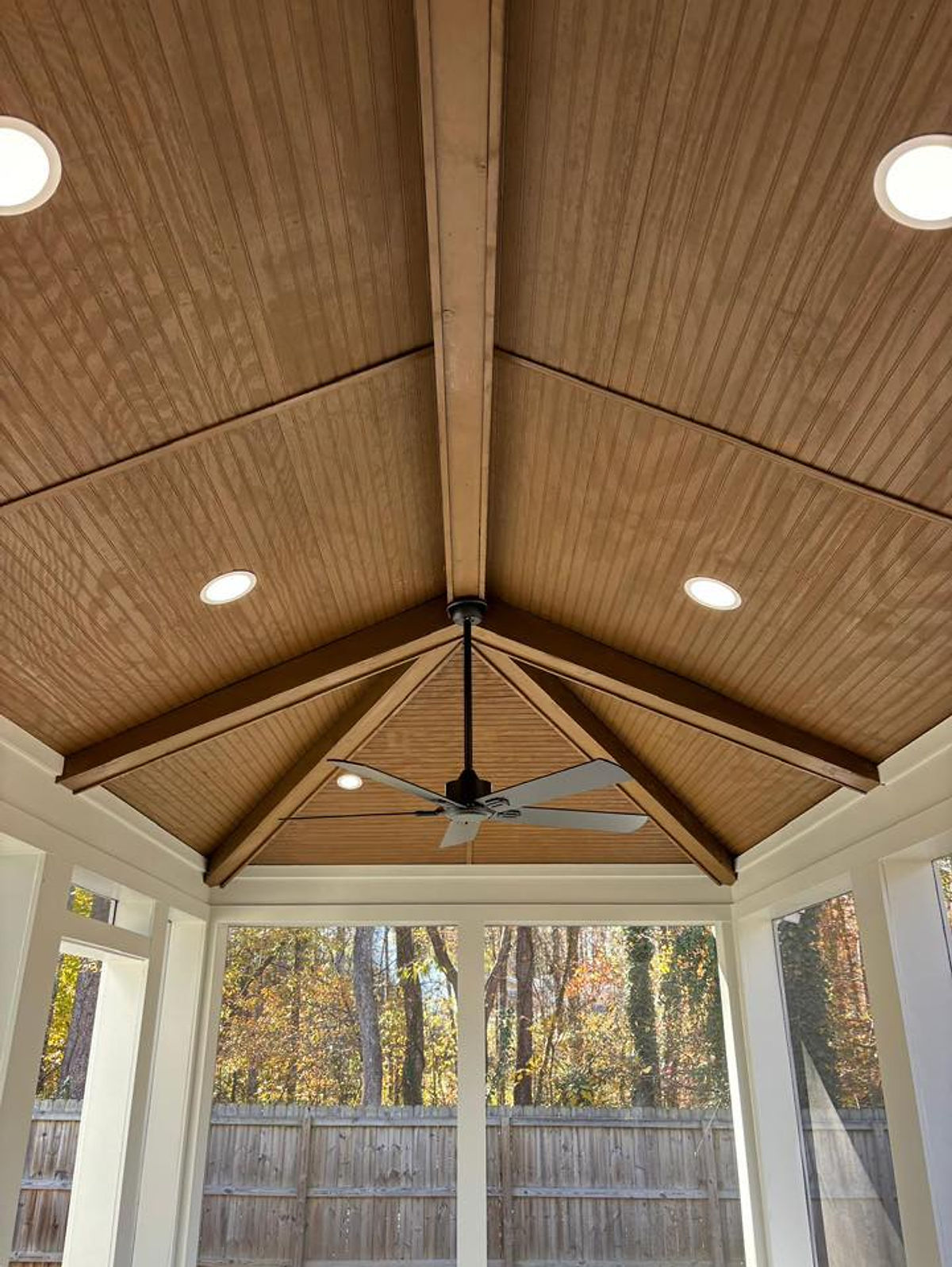 Wooden ceiling with a central fan and recessed lights in a sunroom, overlooking trees and a wooden fence. Bright, cozy atmosphere in porch renovation near Huntersville, NC.
