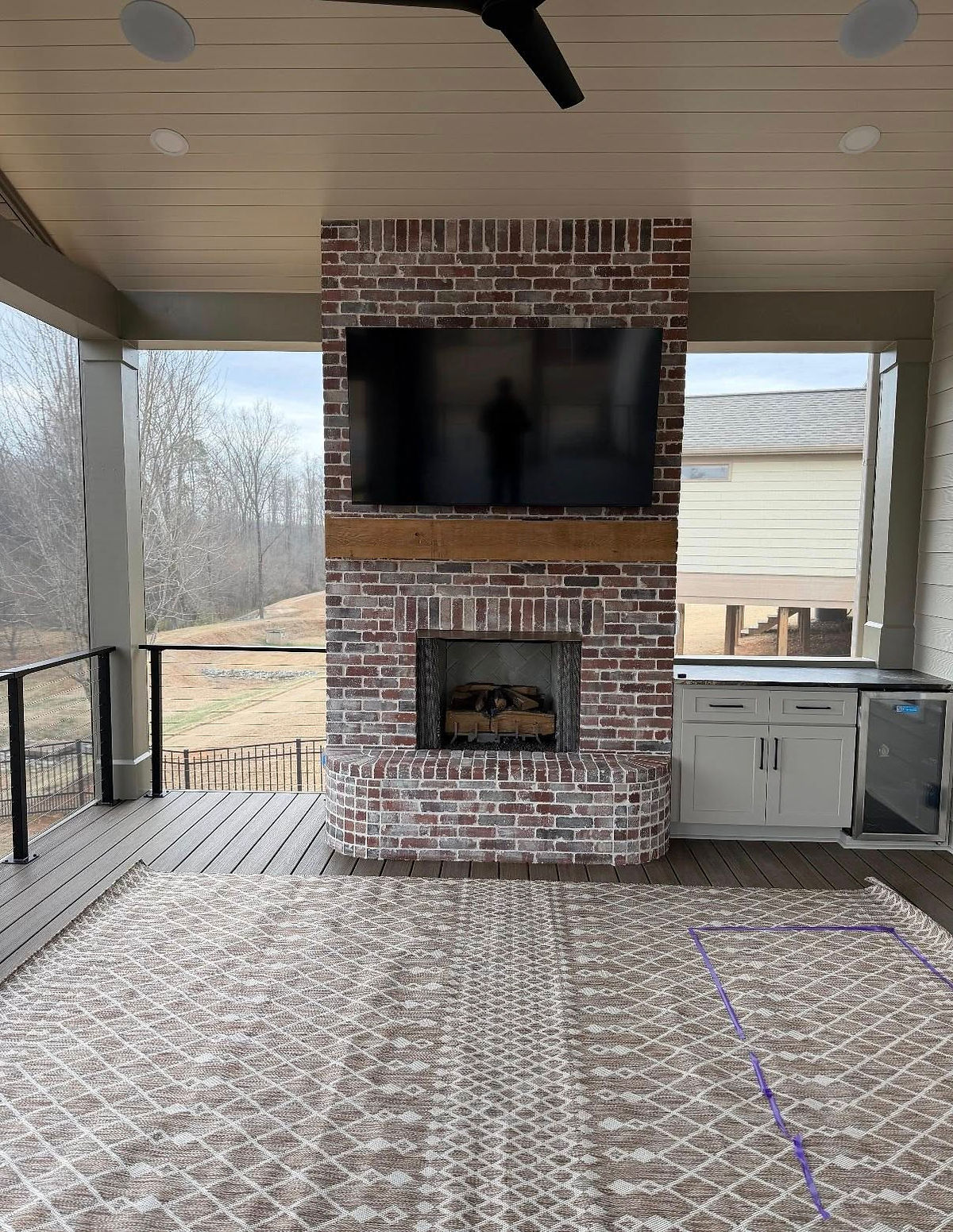 Covered patio with brick fireplace, mounted TV, patterned rug, and outdoor kitchen. Trees visible through open sides, creating a cozy feel for this home renovation  near Cornelius, NC.