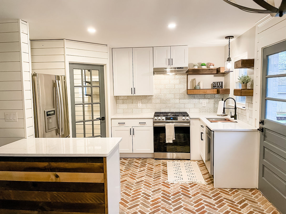 Modern kitchen with white cabinets, stainless steel appliances, and brick-patterned floor. Open shelves hold decor; sign reads 