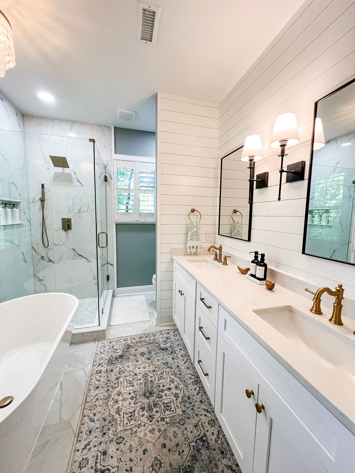 Bright bathroom with marble shower, white vanity, gold fixtures, and a patterned rug. Two mirrors and wall lamps create a cozy ambiance in this home renovation near Cornelius, NC.