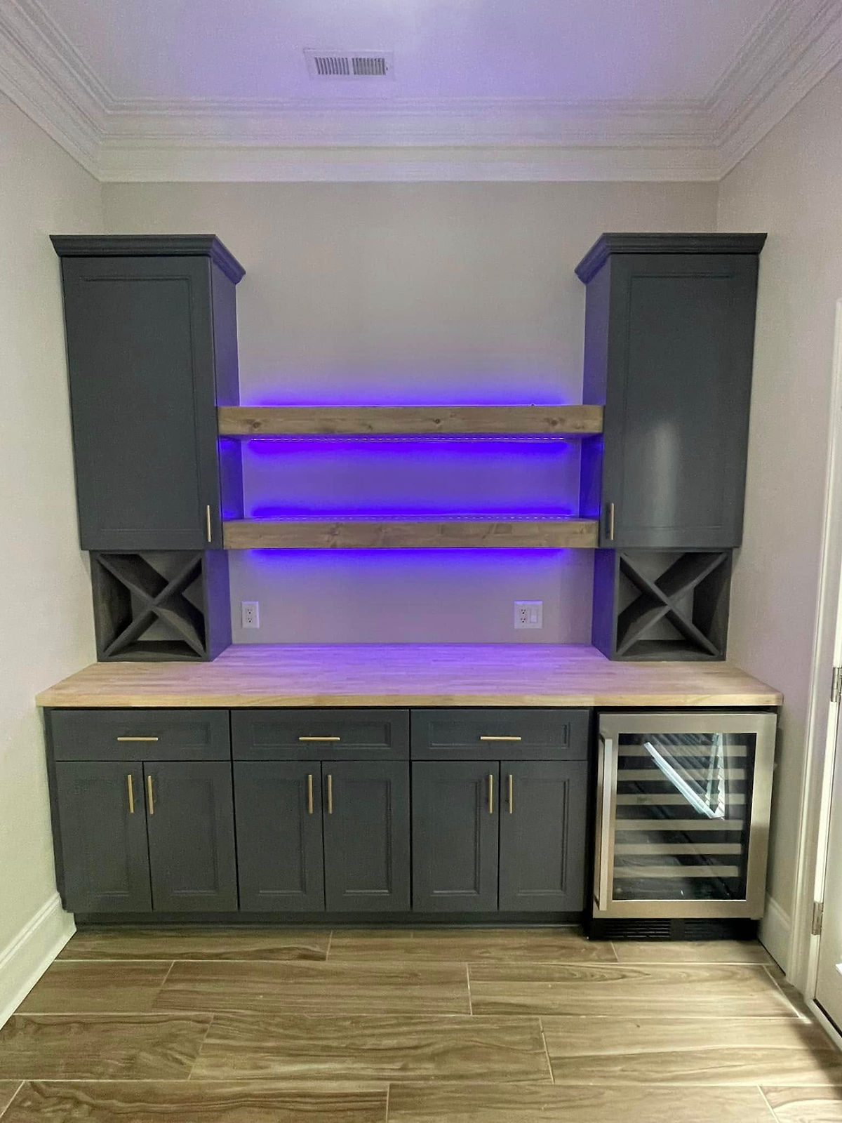 Dark cabinets and a wooden countertop with a wine fridge below purple-lit shelves, set against a beige wall and wooden floor, creating a modern vibe.