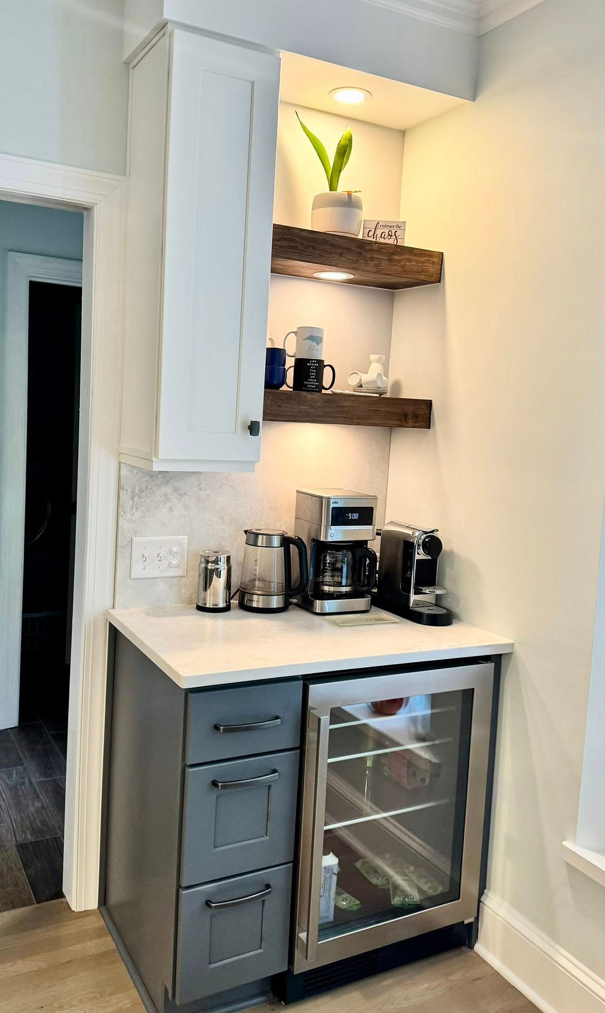 Modern kitchen nook with a coffee station, cabinets, small fridge, and shelves displaying mugs and a plant. Text reads 