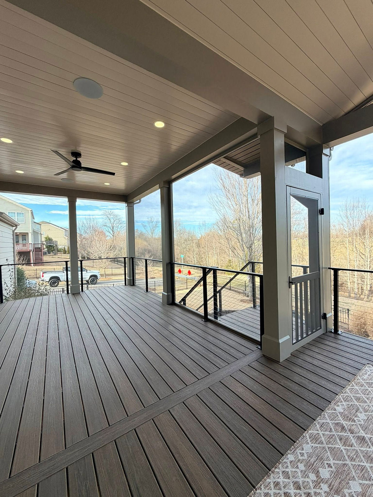 Covered wooden deck with ceiling fan and lights. Overlooks a yard with trees and a parked white truck. Calm and spacious atmosphere.