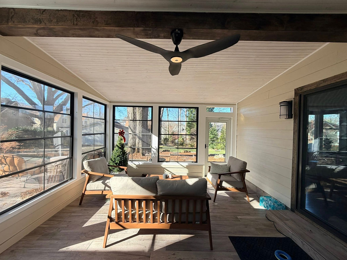 Sunlit enclosed porch with beige seating, a ceiling fan, and large windows overlooking trees. Cozy and inviting atmosphere.