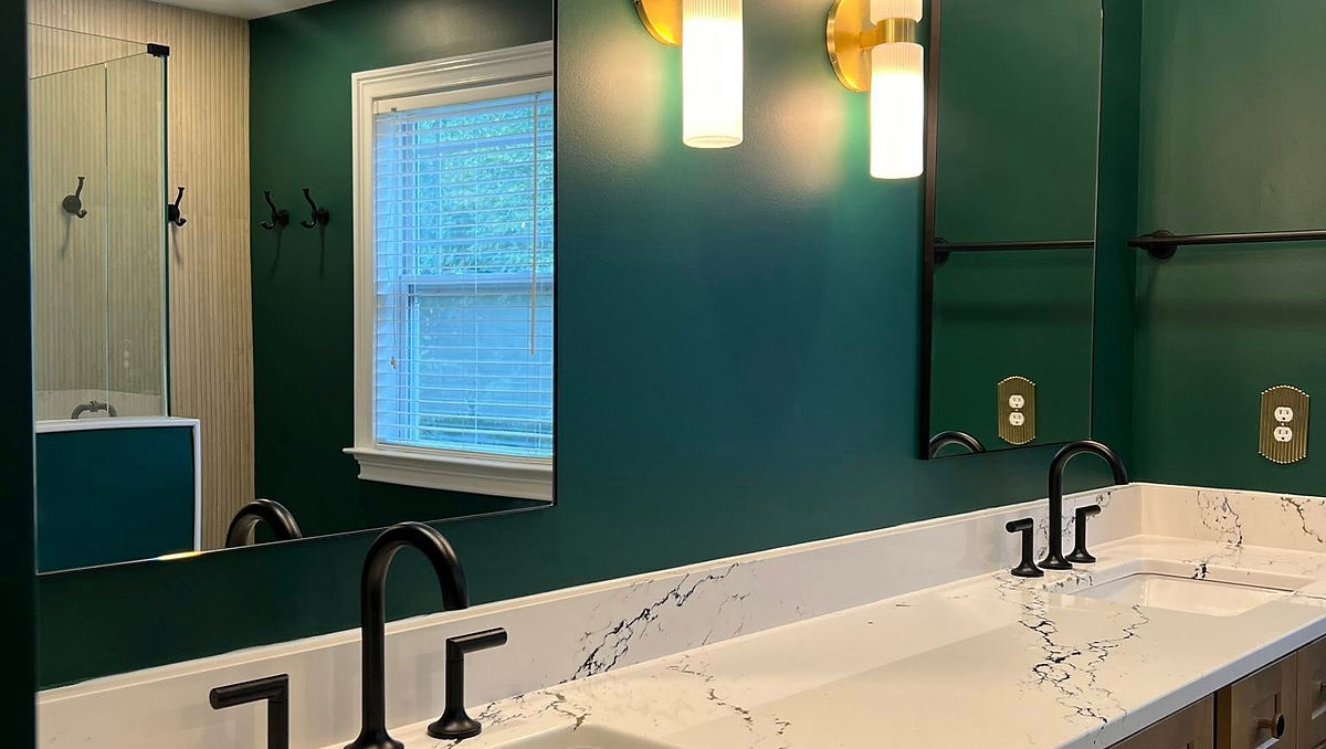 Modern bathroom with dual sinks on a marble countertop, black fixtures, green walls, wooden cabinets, and bright wall lights.