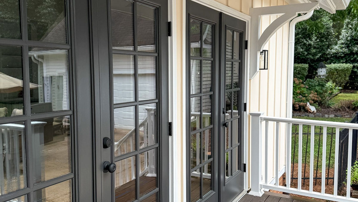 French doors with dark frames on a cream paneled house, open to a wooden deck. Green foliage at the end of the porch visible.