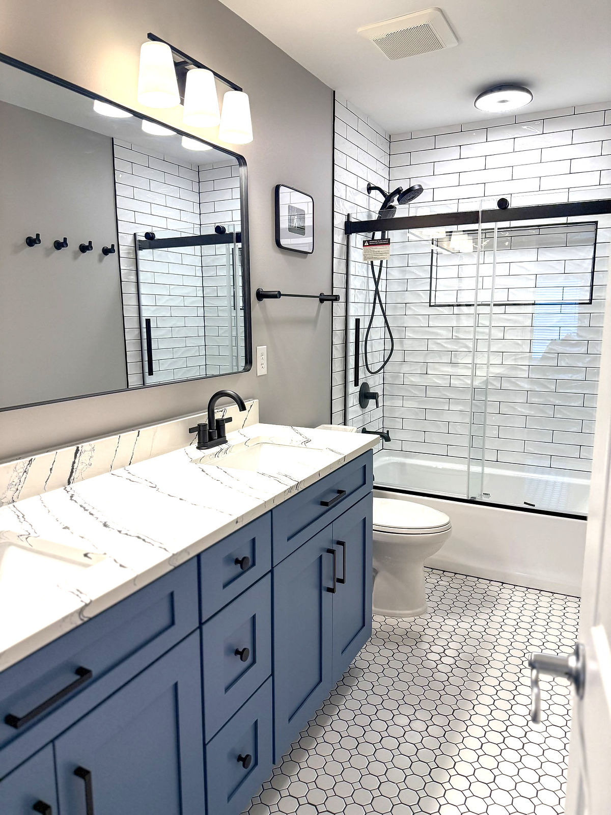 home renovation near Huntersville, NC featuring double sink vanity in blue with black hardware and oversized mirror above. Subway tiled shower with black hardware finishes the back of the room.