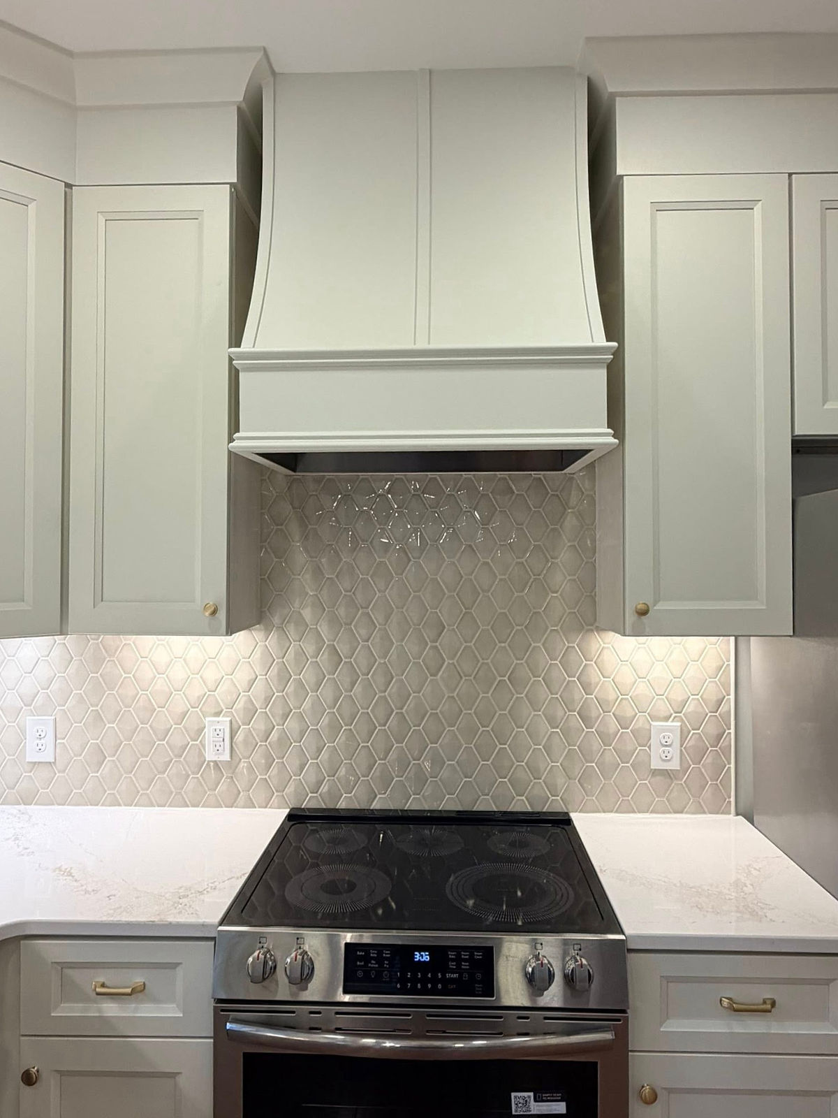 Modern kitchen with light gray cabinets, hexagonal tile backsplash, and an electric stove. Brass handles and soft under-cabinet lighting.