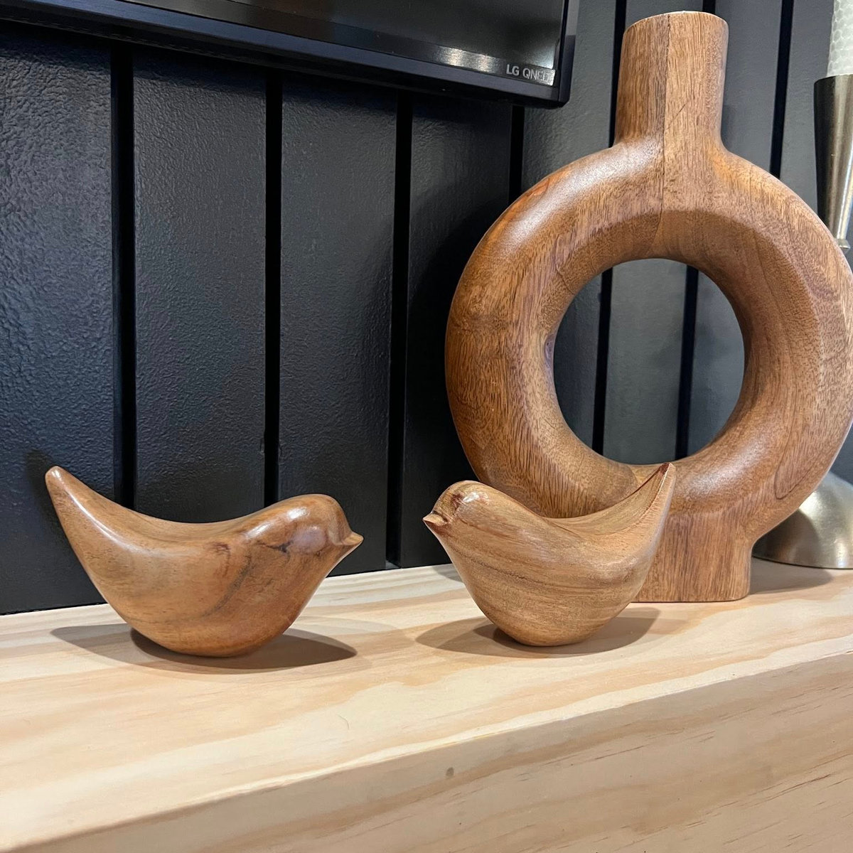 close up of black media wall with wooden mantel. On the mantel are two wooden bird figures in front of a circular wooden sculpture