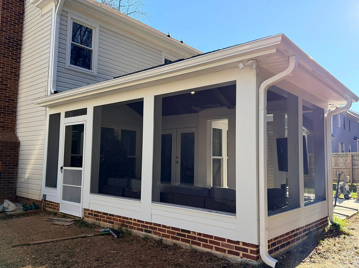 Screened porch attached to a white house with visible doors and windows. Brown brick foundation, yard tools on the ground; sunny day in Huntersville, NC.