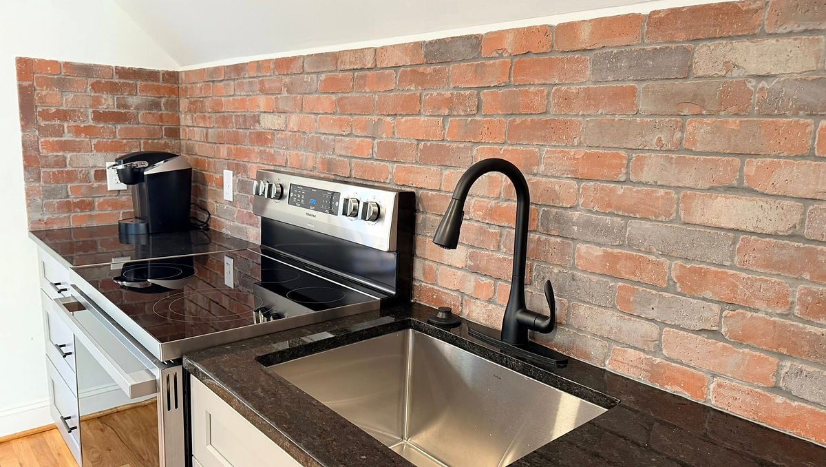 Modern kitchen with brick backsplash, black countertop, stainless steel sink, and appliances. A coffee maker sits beside the stove.