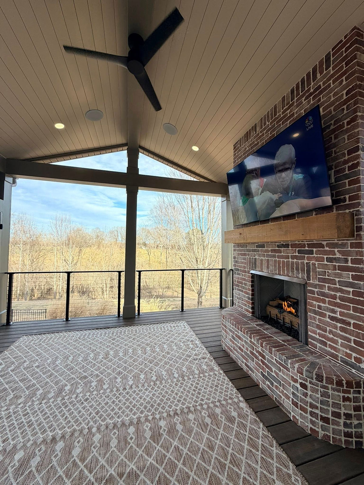 Covered patio with a ceiling fan, brick fireplace, and TV showing sports. Rug with geometric pattern. Scenic view of trees outside.