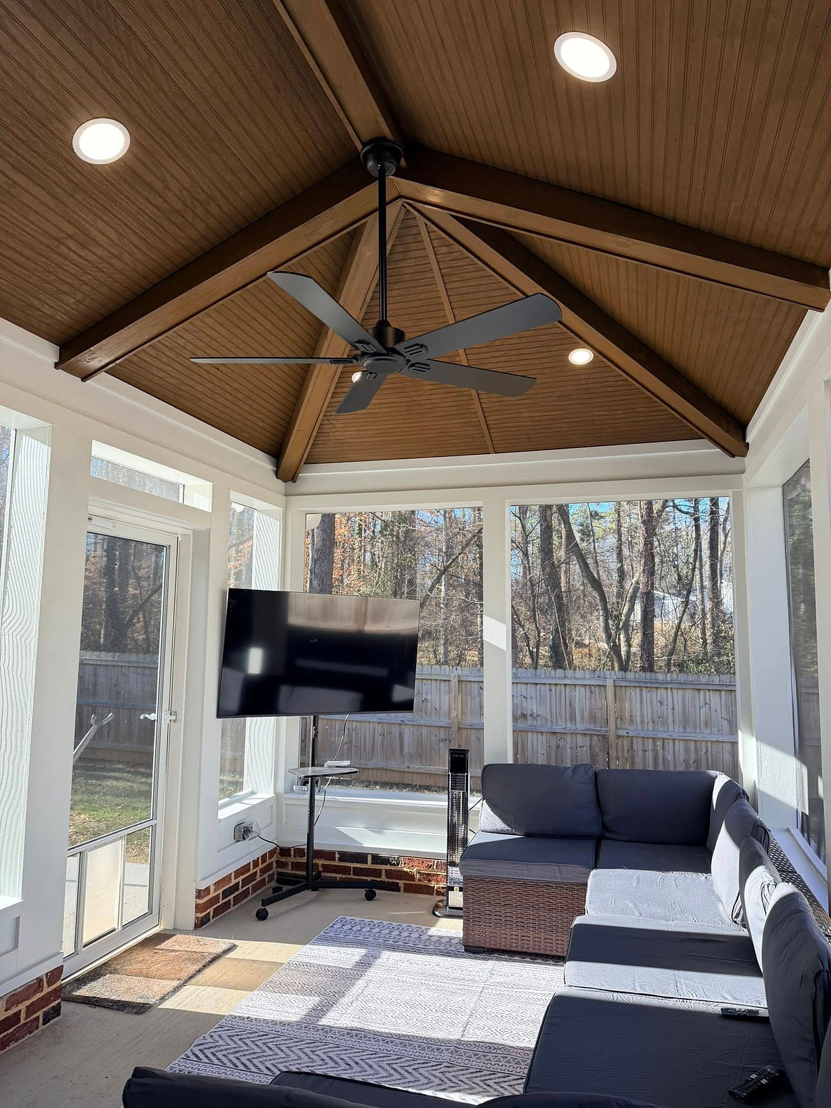 Sunlit porch with gray sectional sofa, wall-mounted TV, and a black ceiling fan. Wooden ceiling and outdoor view through screened windows on this home renovation near Huntersville, NC.