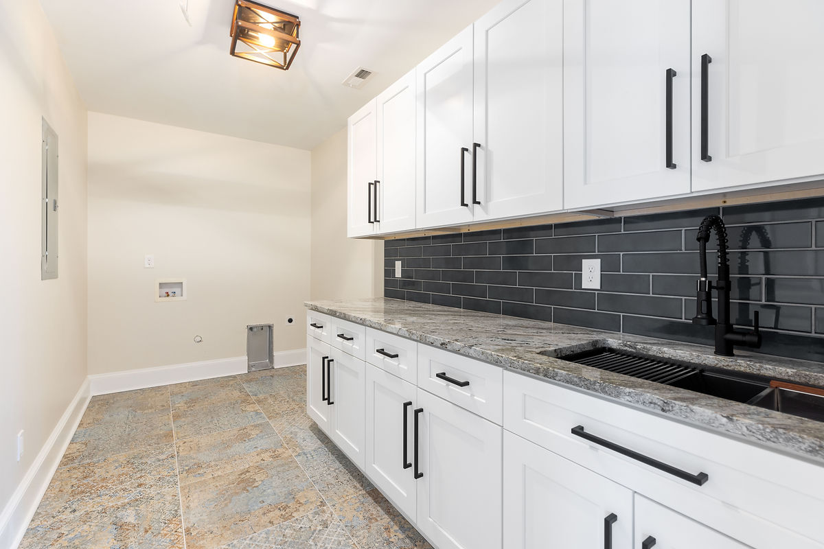 Modern laundry room with white cabinets, dark handles, gray stone countertop, black tile backsplash, and textured tile floor.