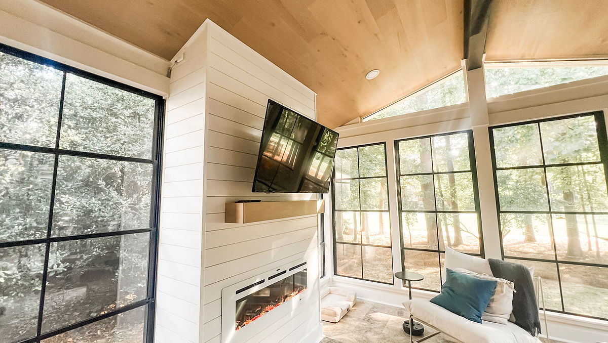 Bright sunroom with large windows, white walls, a wall-mounted TV, and a modern electric fireplace. A cozy chair and pillows enhance the relaxing vibe.