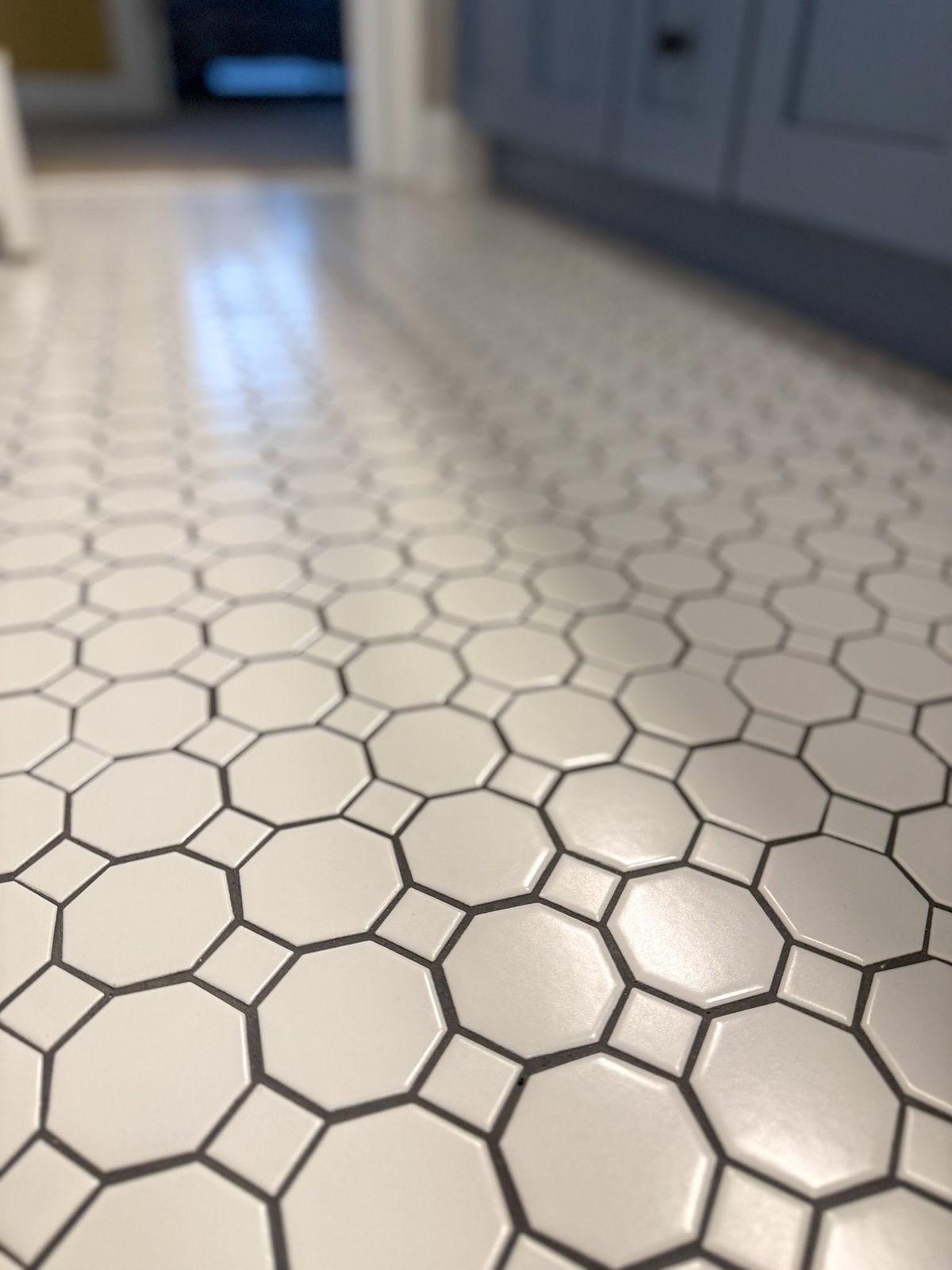 White hexagonal tiles on a floor with gray grout lines, leading to a blurred background of a room with dark and light shades in this home renovation near Cornelius, NC.