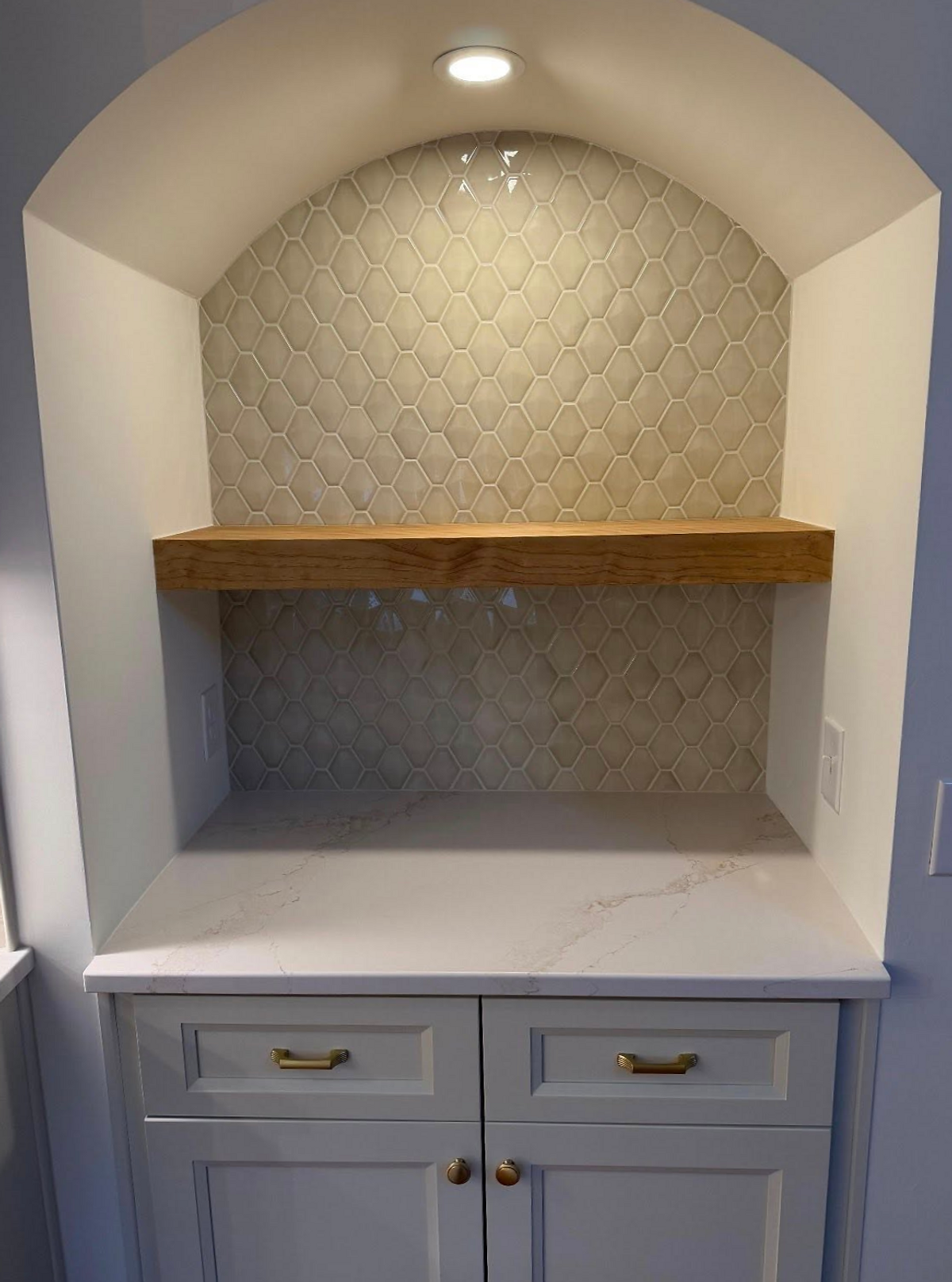 Built-in shelf with wood panel, white cabinets below, and hexagonal tile backsplash. Soft lighting from above creates a cozy ambiance.