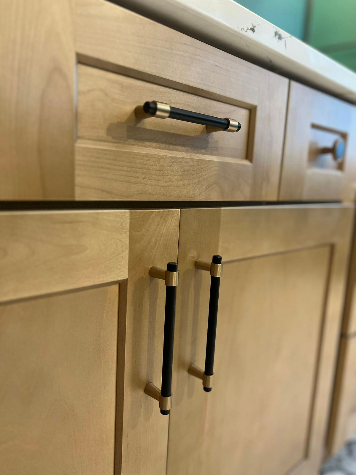 Wooden kitchen cabinets with black and gold handles, set against a white countertop with gray veining. Bright, modern atmosphere.