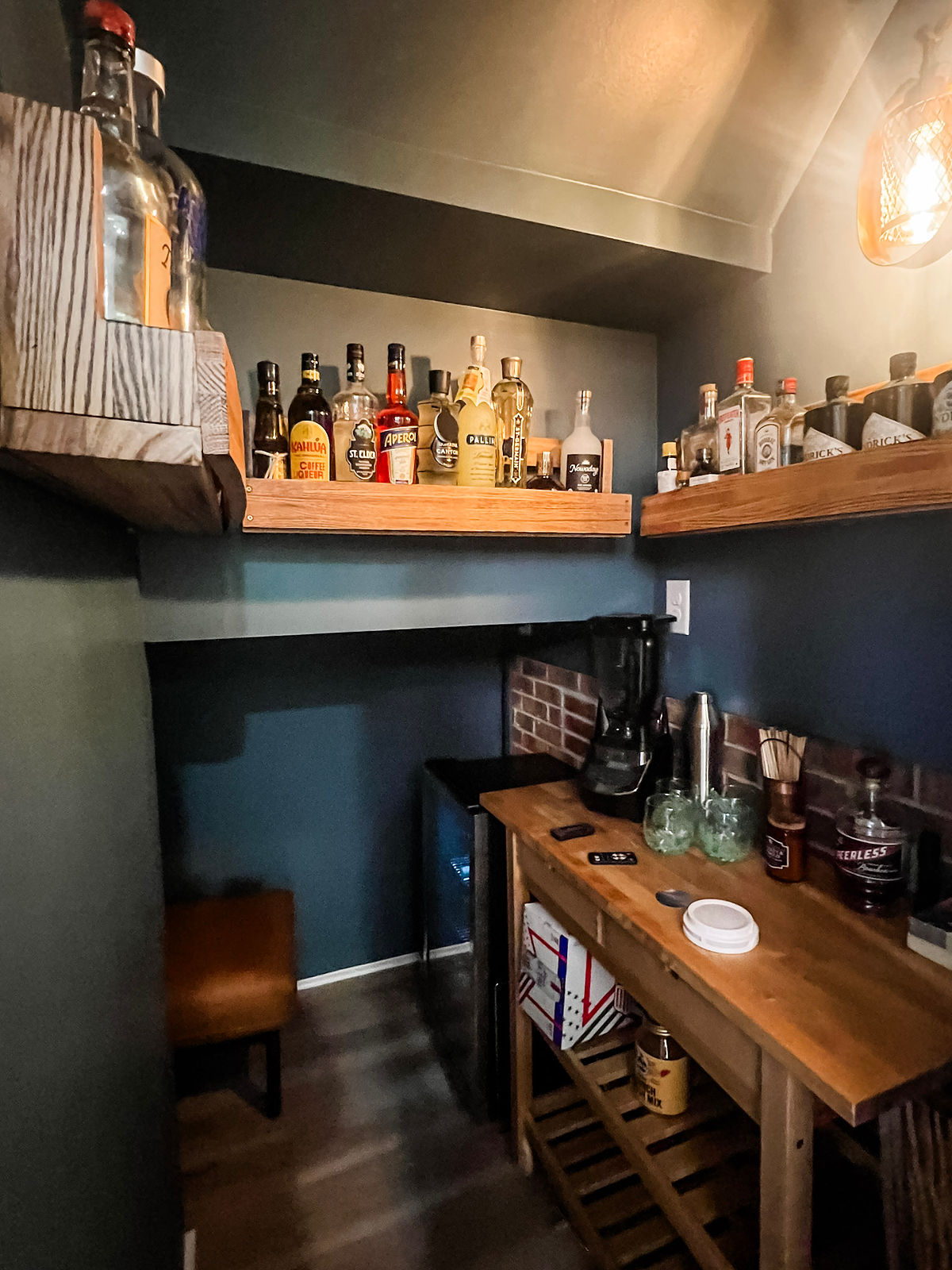 Small home bar with wooden shelves and various liquor bottles. Dark walls, rustic wooden countertop with a blender, and dim lighting. Cozy vibe.