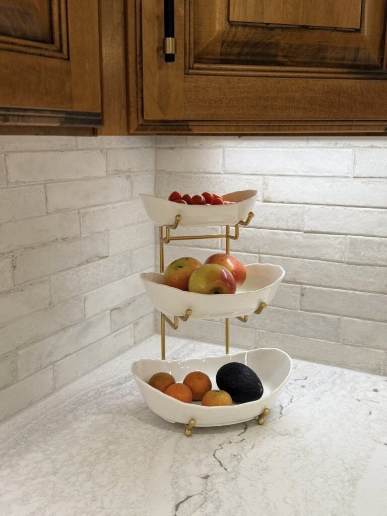 Three tiered white fruit bowl holds fruit on a stone countertop under wood cabinets in a fresh home renovation near Cornelius, NC.