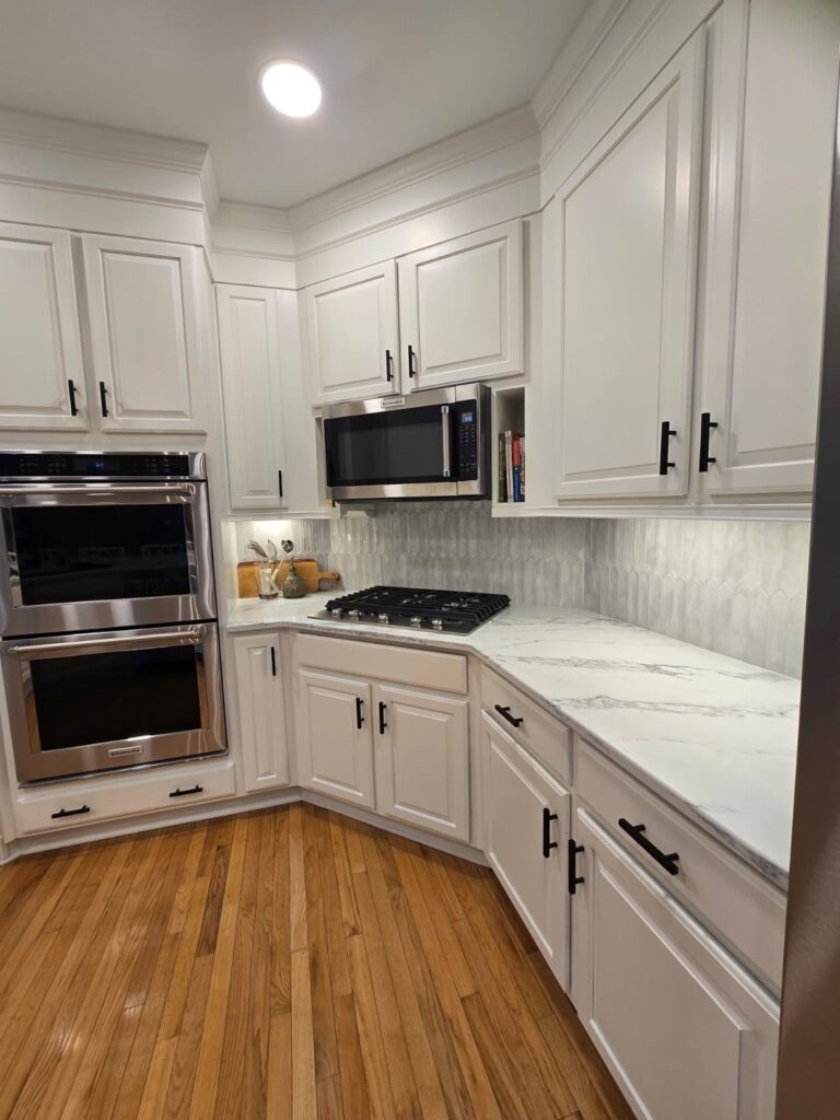 kitchen renovation near Cornelius, NC featuring light stone countertops withe marble backsplash, white cabinetry, wood floors, and stainless appliances.