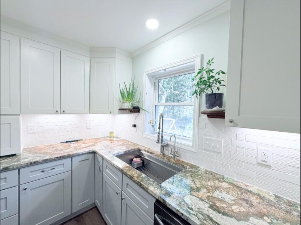 kitchen renovation with a bright interior in cornelius, nc. Features stone counters, white cabinetry, and a sink centered on a window to the right.