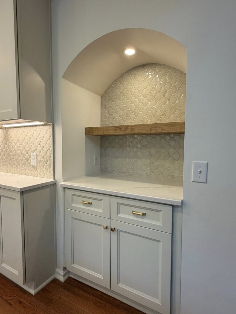 an arched alcove with gray glass tile backsplash, light gray cabinets and open wood shelving