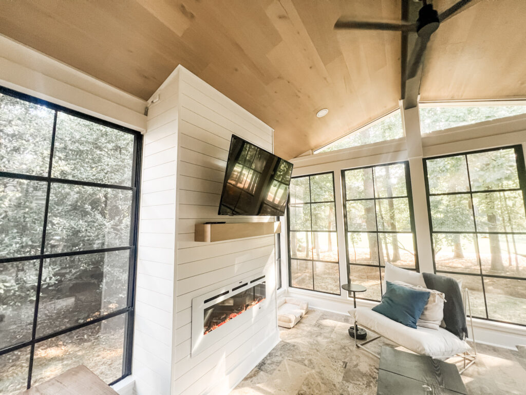 interior of a sun porch near cornelius, NC with stone flooring, floor to ceiling black framed windows and white ship lap walls surrounding a fireplace with v mounted above