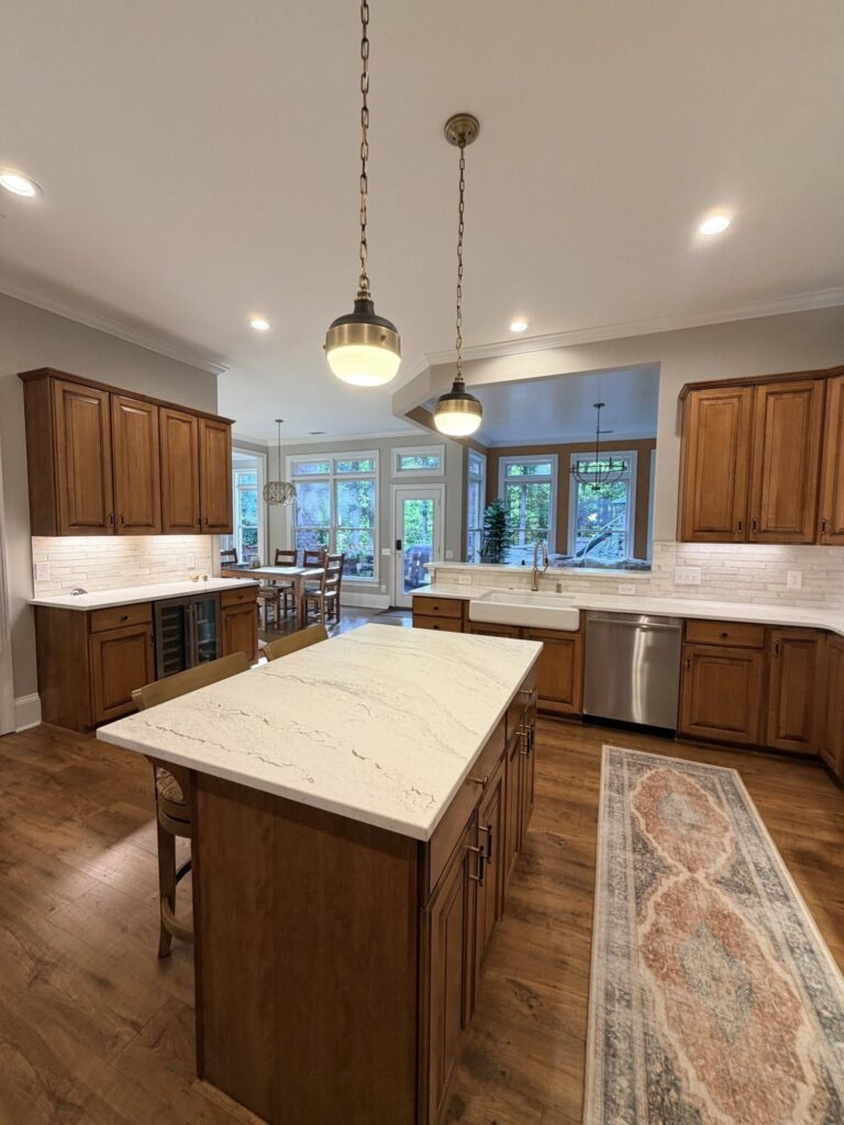 A wide view of a kitchen renovation near Davidson, NC featuring a large stone topped island, wood cabinetry, and metal pendant lights.