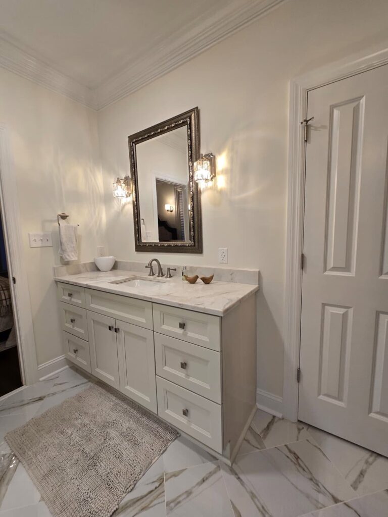 functional luxury renovation near huntersville, nc featuring all white bathroom with marble flooring and cabinetry. Large mirror above sink is framed in ornate metal
