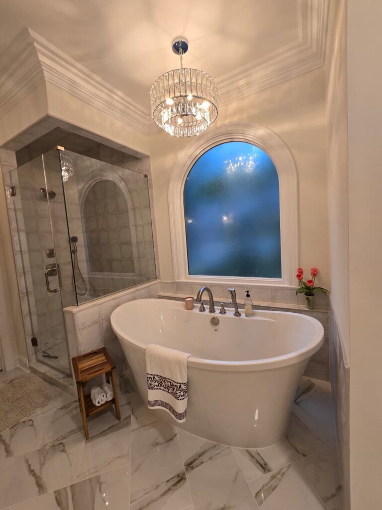 functional luxury renovation of a bathroom. Large crystal chandelier hangs above a soaking tub in front of an arched window and beside a tiled shower.