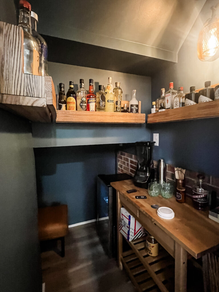 Renovation under a stairway features a moody bar area with shelving, countertop, and exposed brick. An excellent example of home flow design.